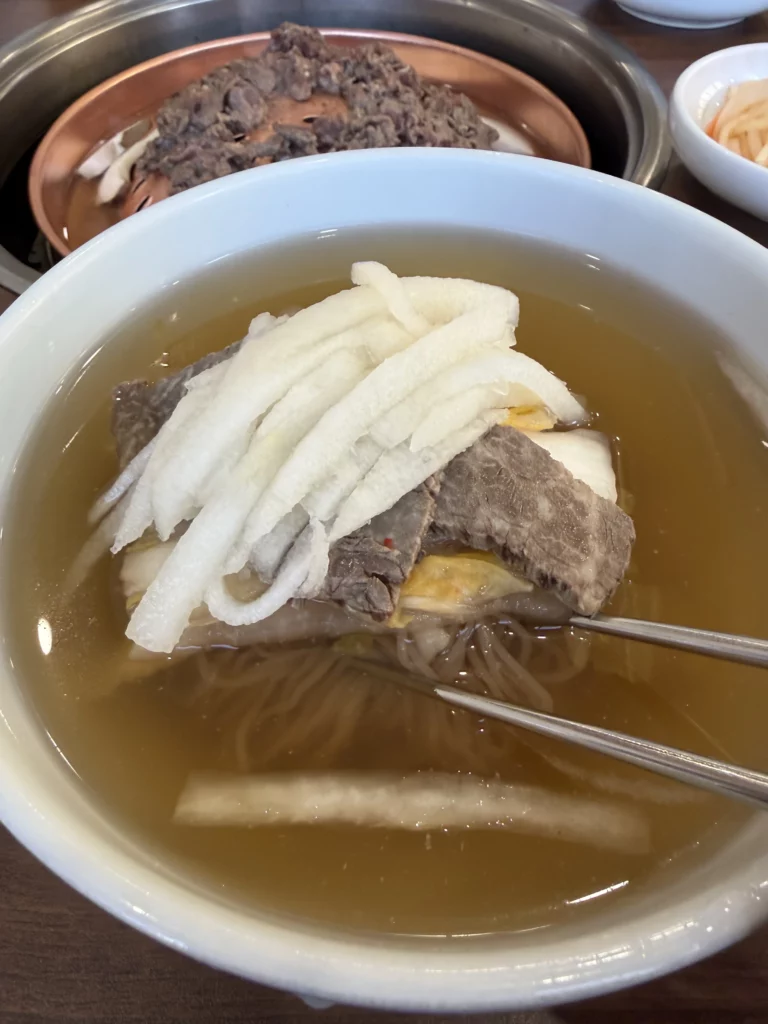 Chopsticks lifting buckwheat noodles from traditional Pyongyang mul naengmyeon at Woo Lae Oak Seoul, showing the delicate texture of the noodles with Korean pear strips, tender beef brisket, and boiled egg in icy beef and dongchimi broth.