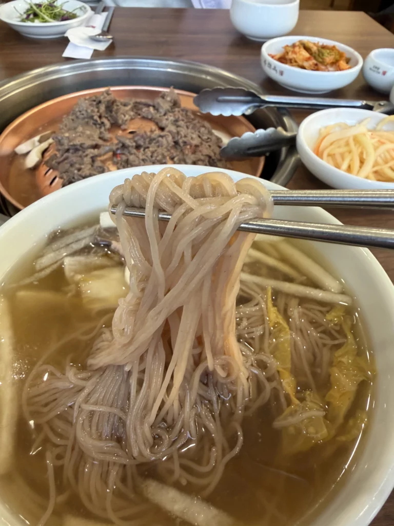 Chopsticks lifting delicate buckwheat noodles from Pyongyang mul naengmyeon at Woo Lae Oak Seoul, demonstrating the traditional eating technique for this authentic Korean cold noodle dish served in beef and dongchimi broth with tableside bulgogi in the background.