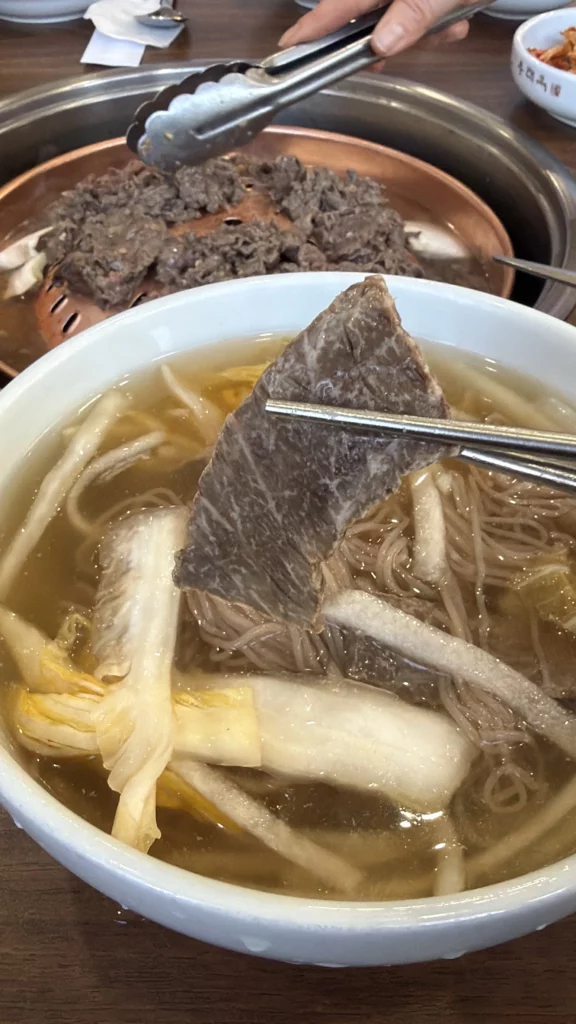 Chopsticks holding tender beef brisket slice from traditional Pyongyang mul naengmyeon at Woo Lae Oak Seoul, revealing the quality meat garnish in the icy beef and dongchimi broth with buckwheat noodles and Korean pear, with tableside bulgogi grilling in the background.