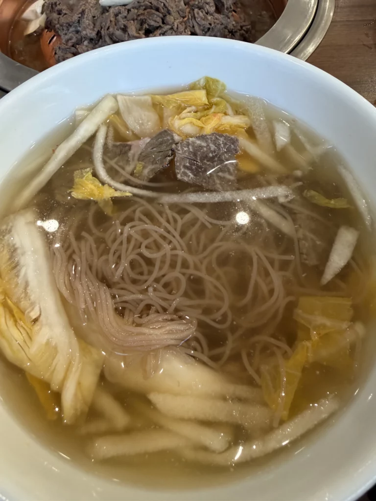 Traditional Pyongyang mul naengmyeon in white bowl at Woo Lae Oak Seoul featuring buckwheat noodles in crystal-clear icy beef and dongchimi broth with Korean pear strips, tender beef brisket, and boiled egg garnishes.