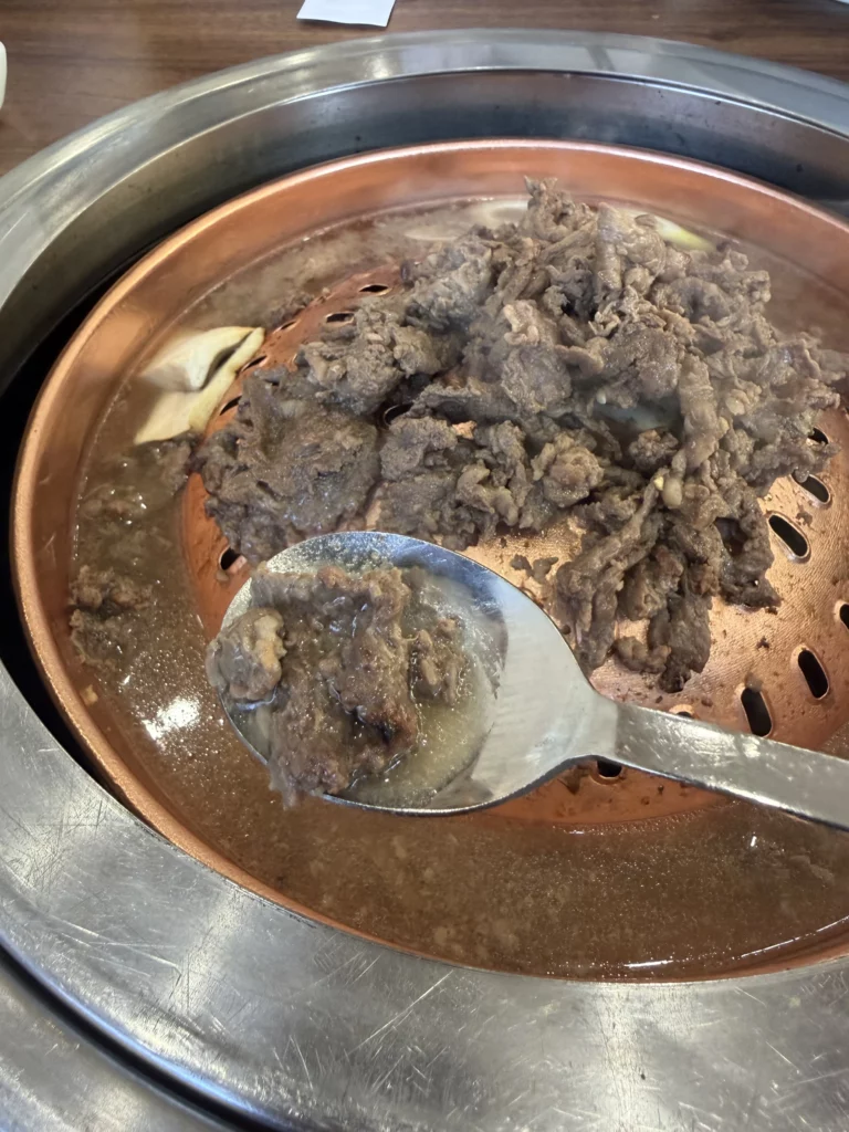 Spoon collecting savory meat juices from grilled Hanwoo beef bulgogi on traditional copper grill at Woo Lae Oak Seoul, showing the caramelized marinated beef with garlic during the tableside Korean BBQ cooking process.