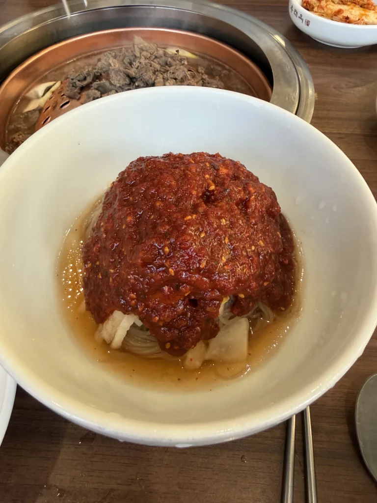 Pyongyang bibim naengmyeon at Woo Lae Oak Seoul showcasing buckwheat noodles covered in vibrant red gochujang sauce with Korean pear and garnishes, served alongside grilling bulgogi in the background.