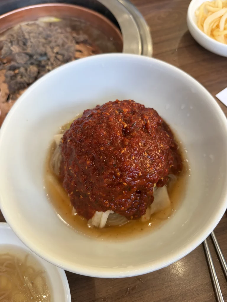 Pyongyang bibim naengmyeon before mixing at Woo Lae Oak Seoul, featuring buckwheat noodles topped with vibrant red gochujang-based sauce and Korean pear strips, served with beef broth for the spicy cold noodle specialty.