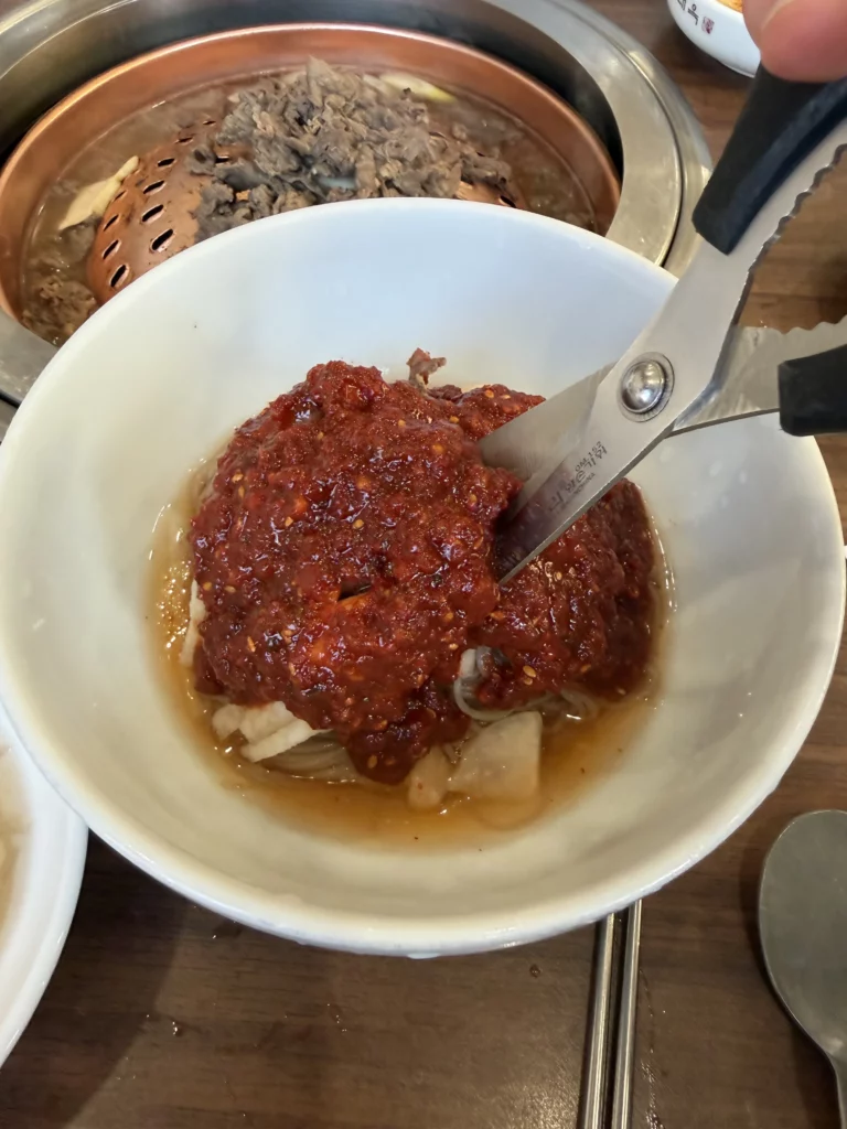 Scissors cutting buckwheat noodles in Pyongyang bibim naengmyeon at Woo Lae Oak Seoul, demonstrating the traditional serving technique to make the spicy gochujang-coated cold noodles easier to eat, with bulgogi grilling in the background.