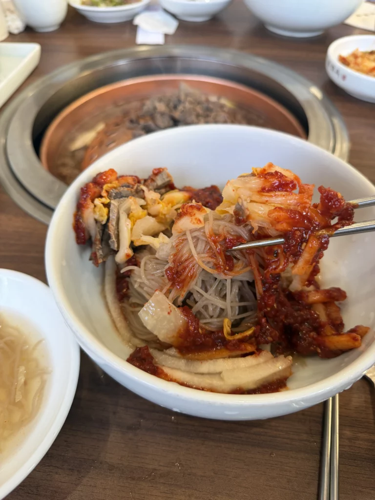 Pyongyang bibim naengmyeon after mixing at Woo Lae Oak Seoul, showing buckwheat noodles thoroughly coated in spicy gochujang sauce with beef, boiled egg, and Korean pear toppings, with copper bulgogi grill visible in the background.