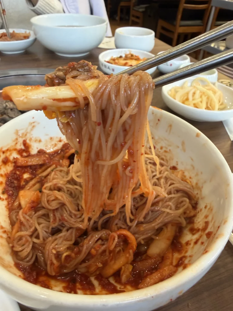 Chopsticks lifting buckwheat noodles coated in spicy gochujang sauce from Pyongyang bibim naengmyeon at Woo Lae Oak Seoul, showcasing the signature red sauce-covered cold noodles with beef and garnishes in this traditional Korean dish.