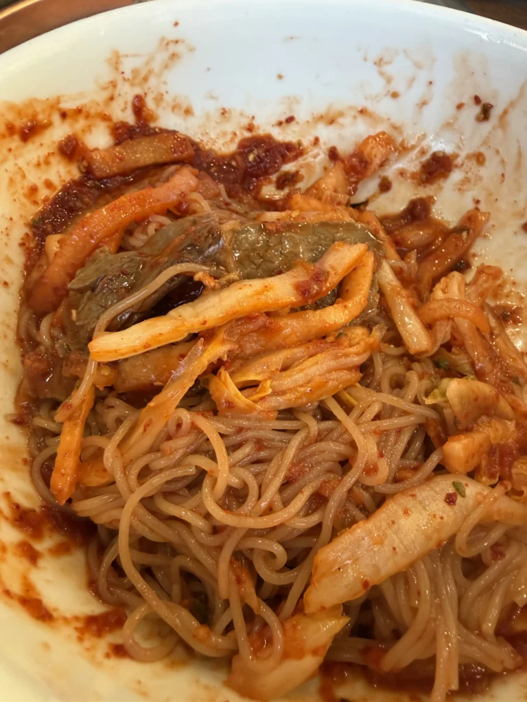Partially eaten Pyongyang bibim naengmyeon at Woo Lae Oak Seoul showing buckwheat noodles thoroughly mixed with spicy gochujang sauce, vegetables, and garnishes after the traditional Korean mixing technique for this cold noodle dish.