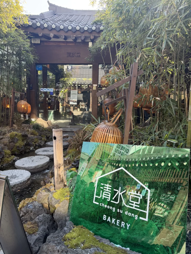 Traditional hanok entrance to Cheongsu-dang Bakery in Seoul's Ikseon-dong neighborhood with bamboo forest, stepping stones crossing a pond, and wooden gate featuring traditional Korean architecture.