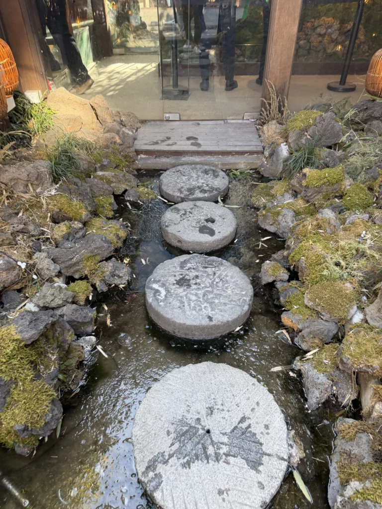 Round stepping stones crossing a serene pond with moss-covered rocks at Cheongsu-dang Bakery's iconic entrance in Ikseon-dong Seoul, leading visitors to the traditional hanok cafe.