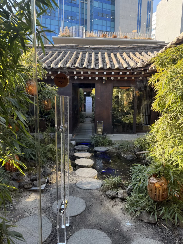 Traditional hanok entrance gate with curved roof tiles at Cheongsu-dang Bakery in Ikseon-dong Seoul, featuring stepping stone pathway through bamboo garden with modern skyscraper backdrop.