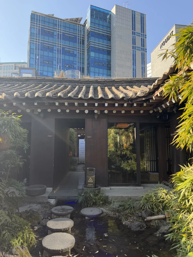 Traditional hanok entrance gate with curved Korean roof tiles at Cheongsu-dang Bakery in Ikseon-dong Seoul, featuring stepping stones across a pond with modern skyscrapers in the background.