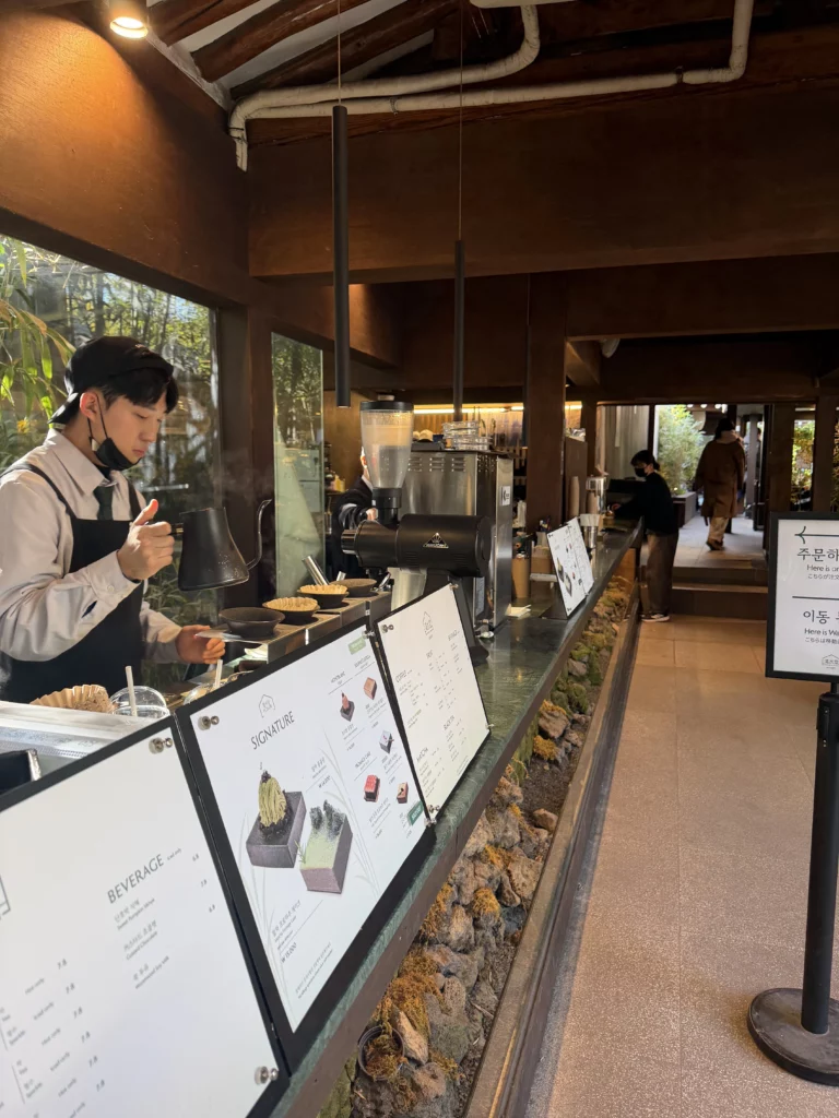 Ordering counter with menu boards at Cheongsu-dang Bakery featuring moss-lined water features, traditional hanok wooden beams, and bamboo garden views at this Seoul Ikseon-dong cafe famous for souffle castella.