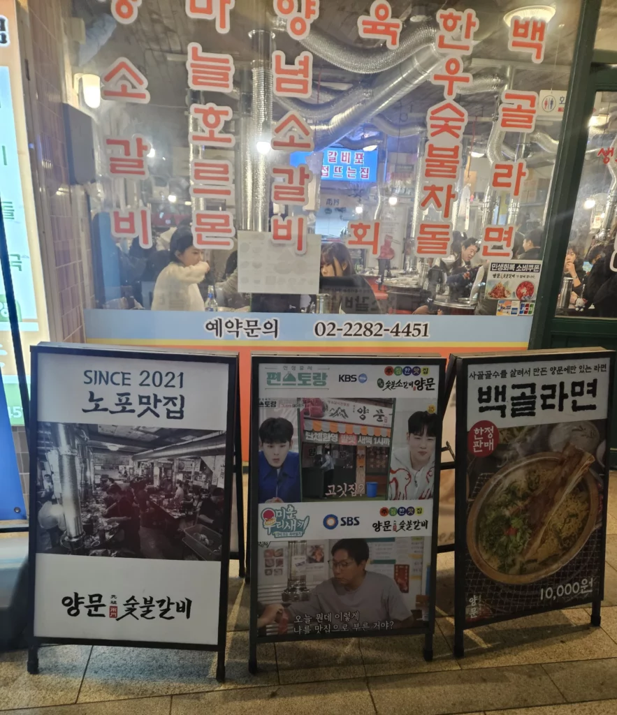 Crowded Korean BBQ restaurant interior seen through glass windows, with customers enjoying grilled beef Korean BBQ and traditional charcoal grilling.