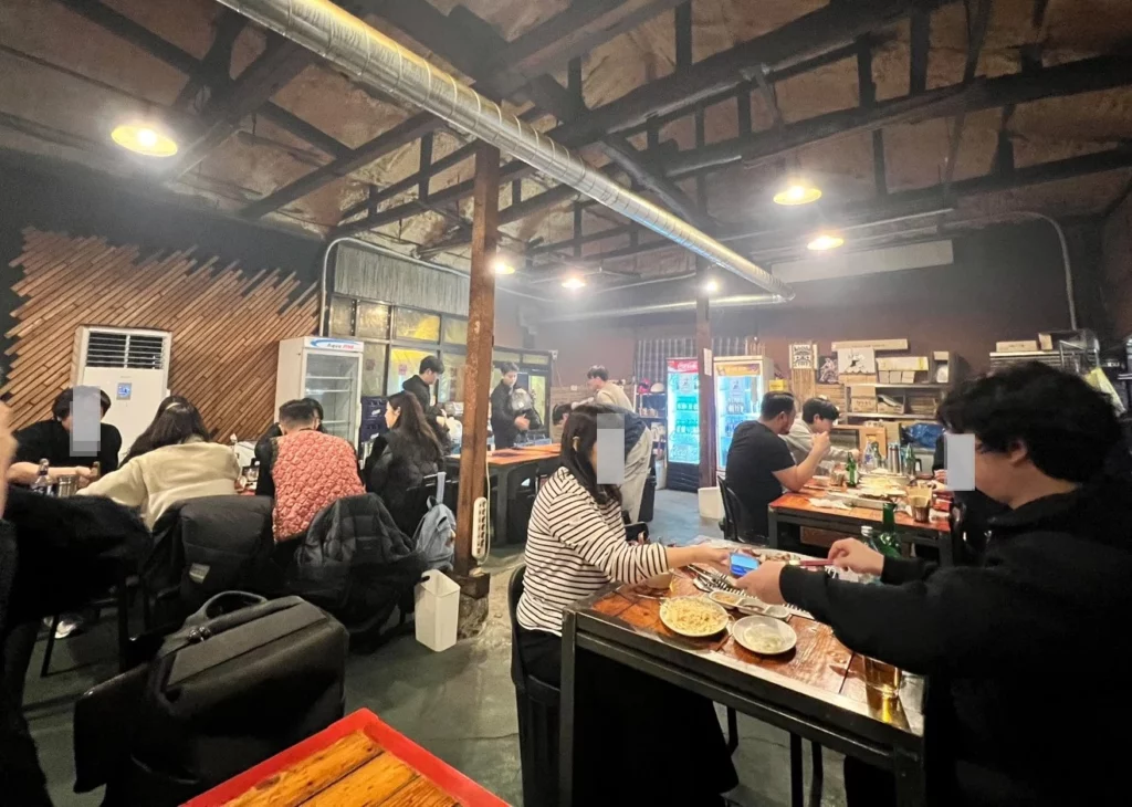 Lively interior of a local Yongsan smoked K-BBQ restaurant, filled with diners enjoying charcoal-grilled meats in a rustic, authentic Korean barbecue atmosphere.