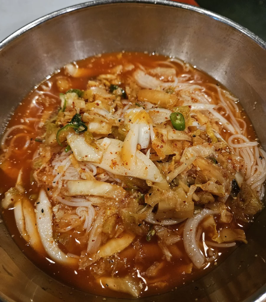 Spicy Korean cold noodles served in a metal bowl at a Yongsan restaurant, topped with kimchi, vegetables, and chili seasoning as a refreshing side dish to rich smoked K-BBQ.