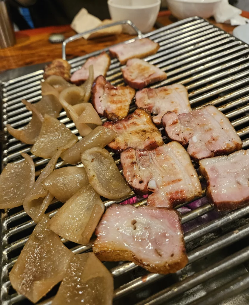 Smoked pork slices and radish grilling together on a metal grate in Yongsan, highlighting traditional smoked K-BBQ with rich aroma, caramelized edges, and classic Korean BBQ style.