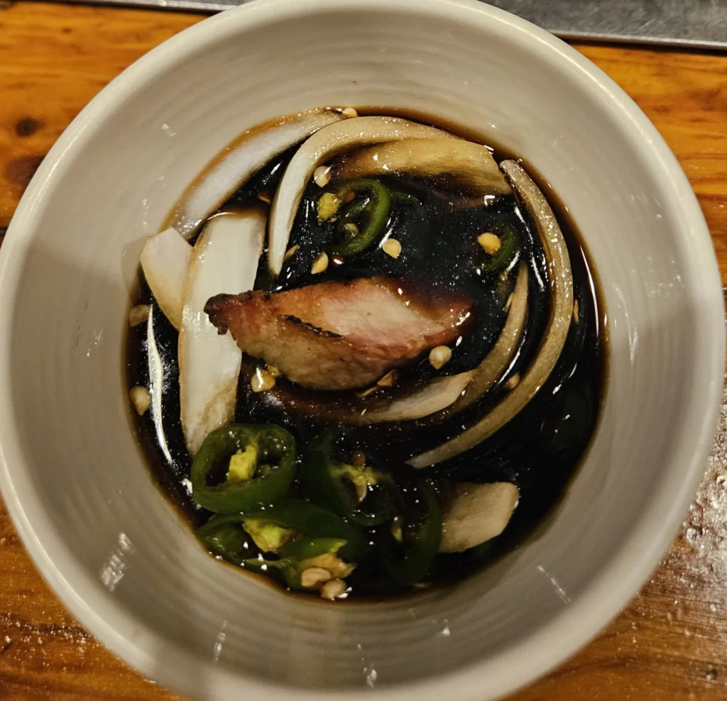 Soy-based dipping sauce with grilled pork, sliced onions, and green chili peppers served at a Yongsan restaurant, complementing the smoky richness of authentic smoked K-BBQ.