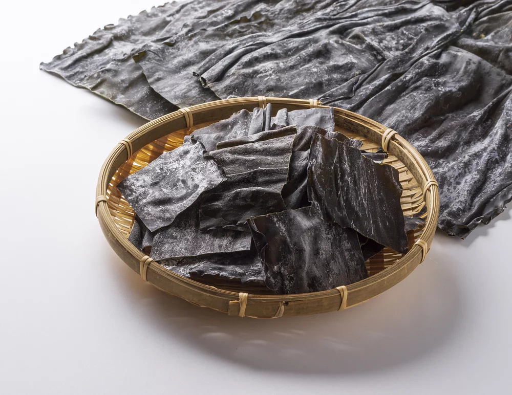 Dried Korean kelp dashima pieces in a bamboo basket with large whole sheet displayed showing white mineral coating and umami-rich surface.