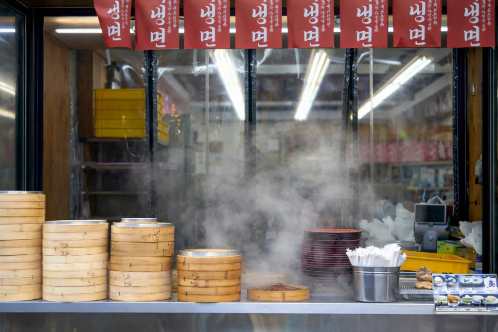 Bamboo steamers stacked high releasing steam at a Bukchon Son Mandu kitchen with naengmyeon signs displayed above.