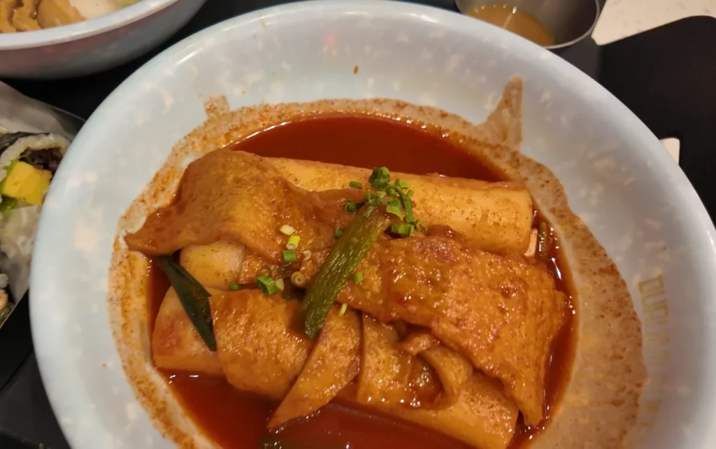 Durumi Bunsik tteokbokki bowl with thick rice cakes and fish cake pieces coated in glossy gochujang sauce topped with green onion.