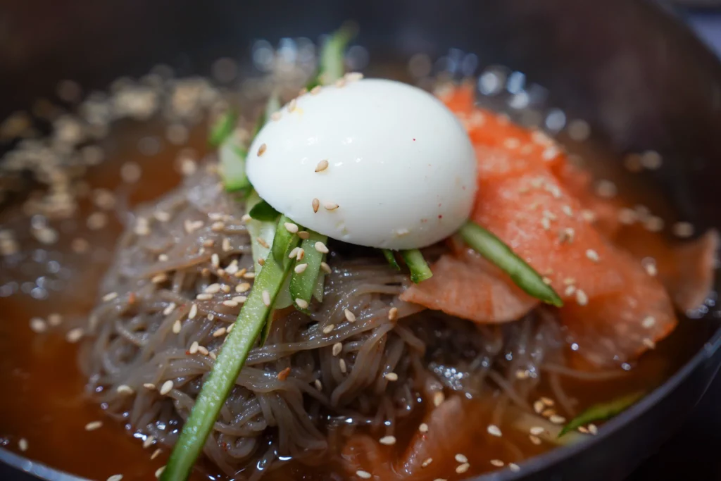 Bukchon Son Mandu cold buckwheat naengmyeon noodles in kimchi broth topped with egg, sesame seeds, and cucumber.