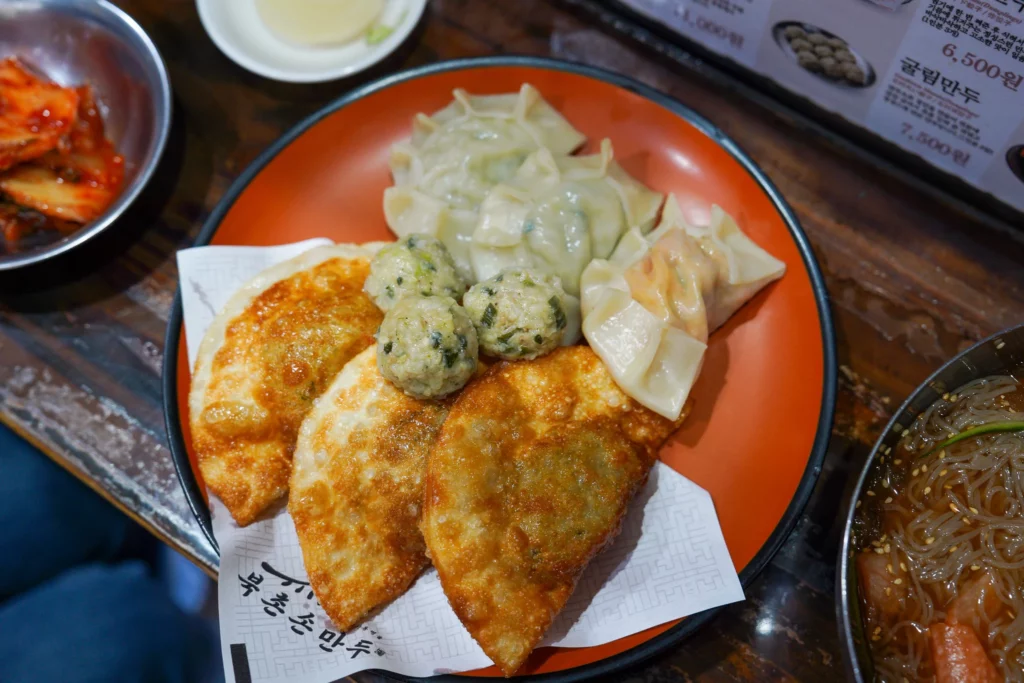Bukchon Son Mandu assorted mandu platter with deep-fried dumplings, steamed mandu, and gullin mandu balls served on an orange plate with kimchi side dish.