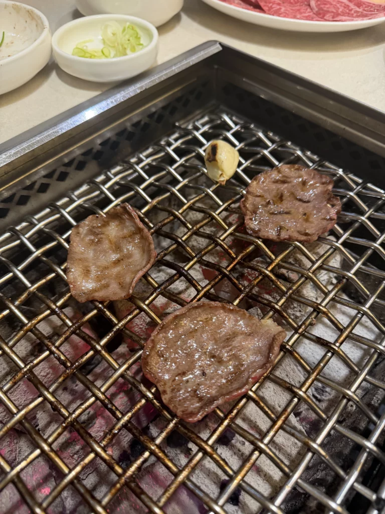 Perfectly grilled beef tongue slices with golden caramelized surface and roasted garlic on charcoal grill at Chowon Korean BBQ in Gangnam Seoul.