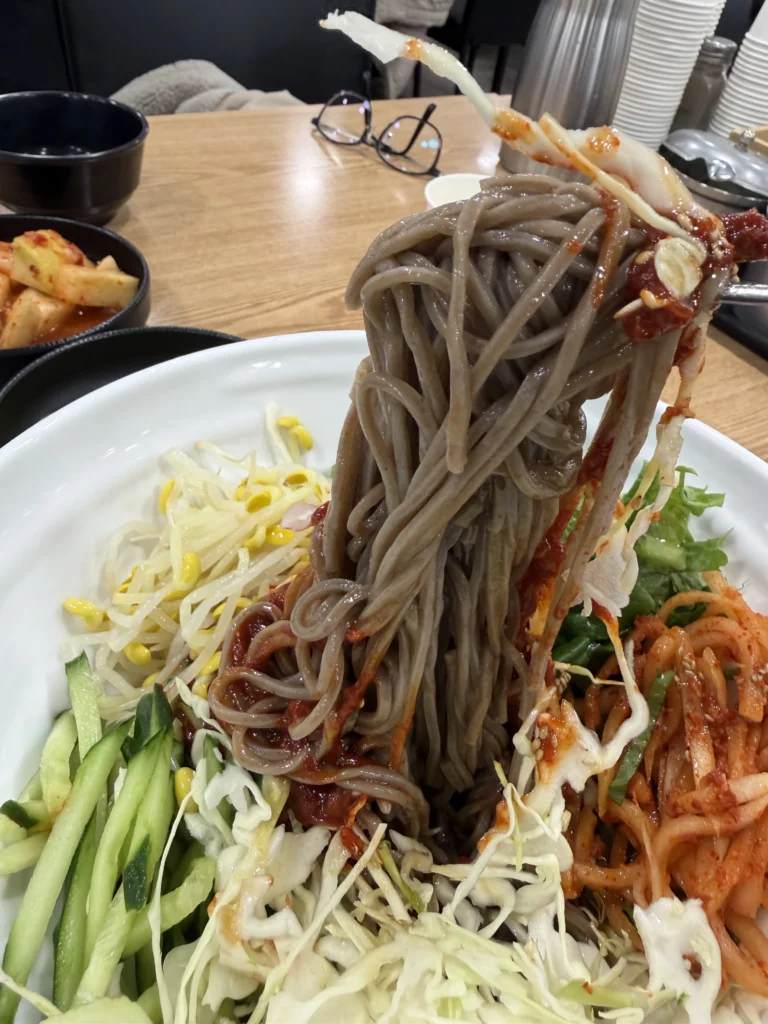 Chopsticks lifting bibim memil myeon spicy buckwheat noodles with gochujang sauce at Mijin Michelin-starred Korean restaurant showing traditional jjolgit chewy texture.