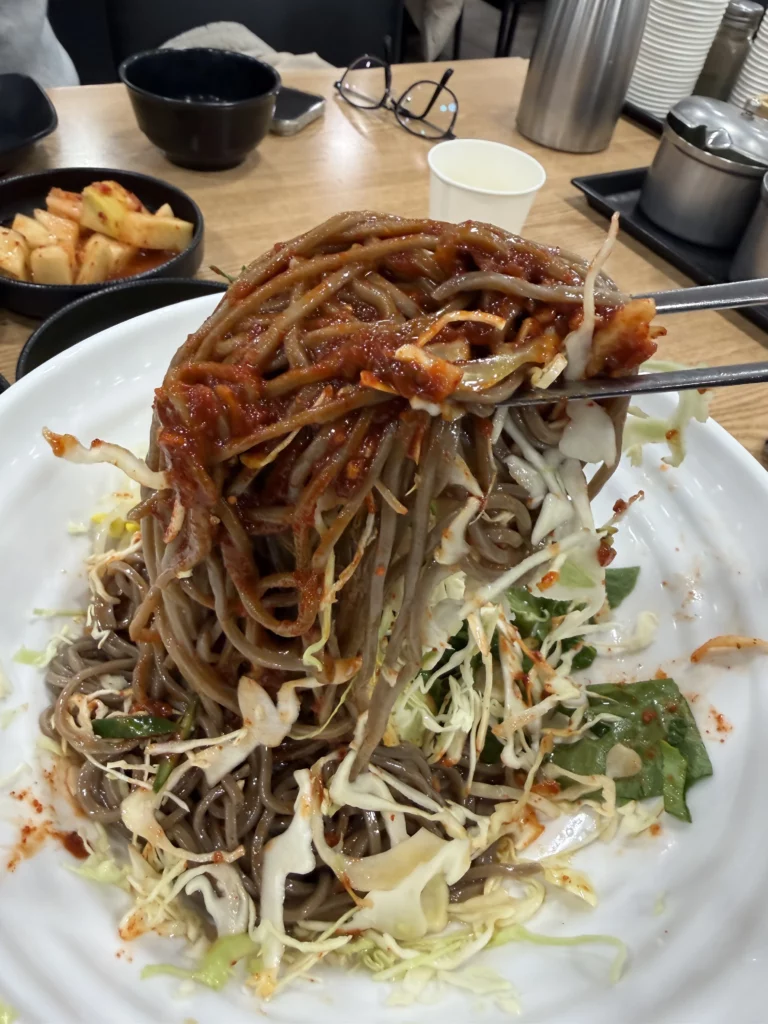 Mixed bibim memil myeon spicy buckwheat noodles with gochujang sauce being lifted with chopsticks at Mijin Michelin Bib Gourmand Korean restaurant in Seoul.