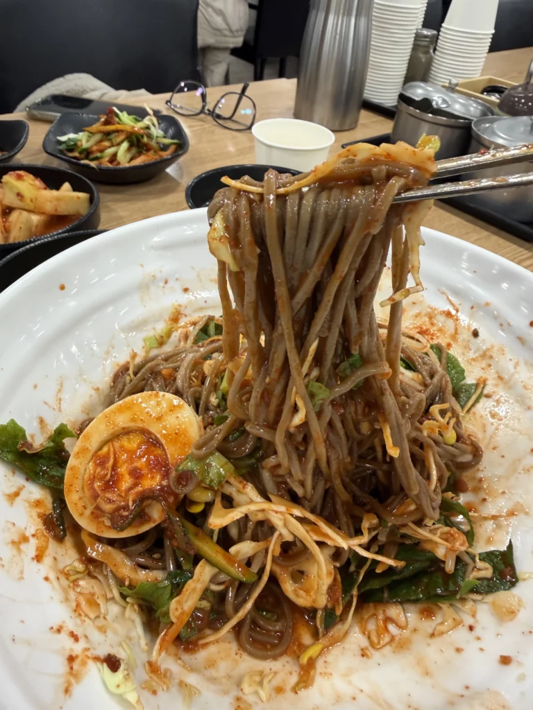 Bibim memil myeon Korean buckwheat noodles being lifted with chopsticks showing soft-boiled egg and spicy gochujang sauce at Mijin Michelin Bib Gourmand restaurant in Seoul.