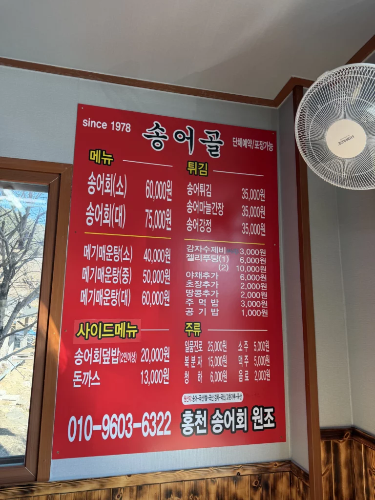 Wall-mounted menu board at 송어골 (Songeogol) showing Korean trout sashimi (송어회) prices — small 60,000 won and large 75,000 won — alongside maeuntang, fried trout, and side dishes, with the restaurant's phone number and 