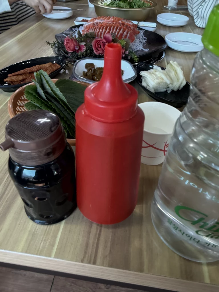 Table setting at Vivaldi Songeogol showing a choganjang (초장) squeeze bottle and soy sauce alongside fresh perilla ssam leaves, with a Korean trout sashimi (송어회) platter visible in the background.