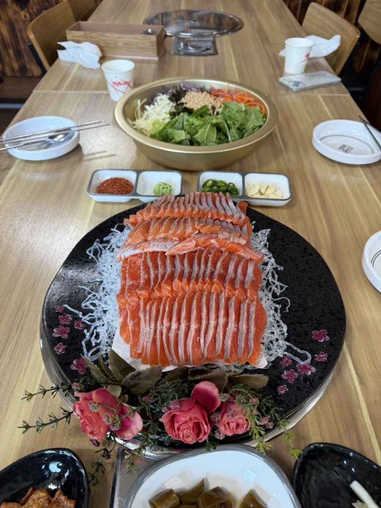 A generous serving of Korean trout sashimi (송어회) set at Vivaldi Songeogol, with thinly sliced fresh trout stacked on shredded radish, choganjang, wasabi, sliced chili, garlic, and a large vegetable salad bowl — the complete freshwater fish dining experience in Hongcheon, Gangwon Province.
