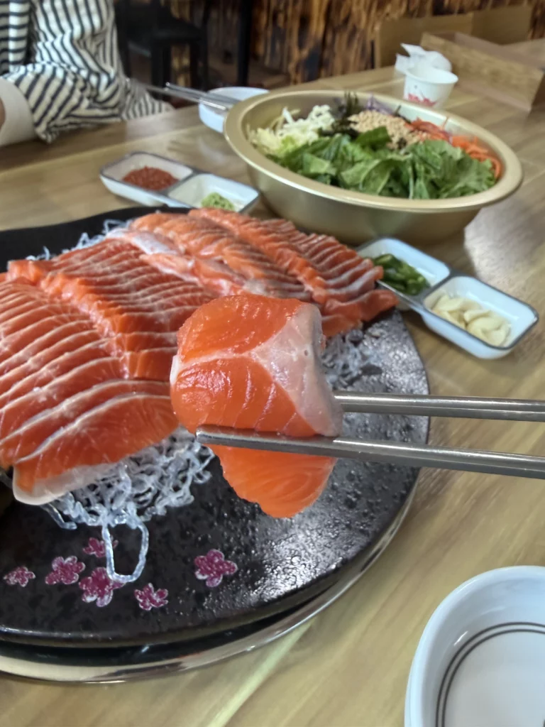 Chopsticks lifting a thick, glistening slice of fresh Korean trout sashimi (송어회) from the platter at Vivaldi Songeogol, with the full set of accompaniments — vegetable salad, dipping sauces, and garnishes — visible in the background.