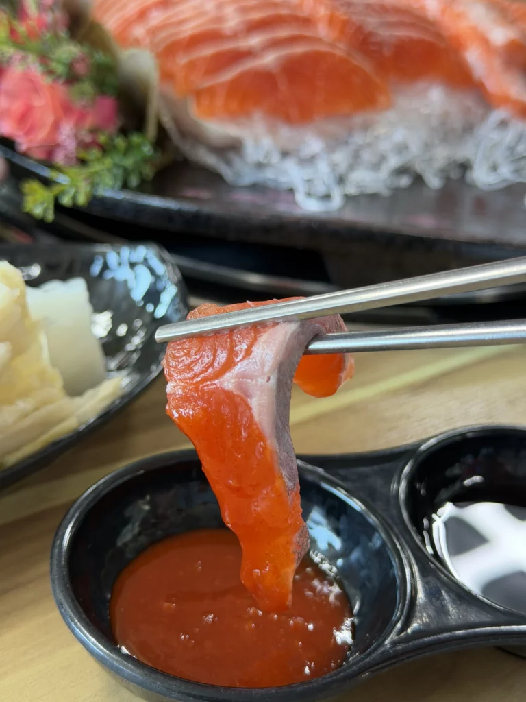 Chopsticks dipping a fresh slice of trout sashimi (송어회) into choganjang (초장) — the classic first step of eating Korean freshwater fish the traditional way at Vivaldi Songeogol.