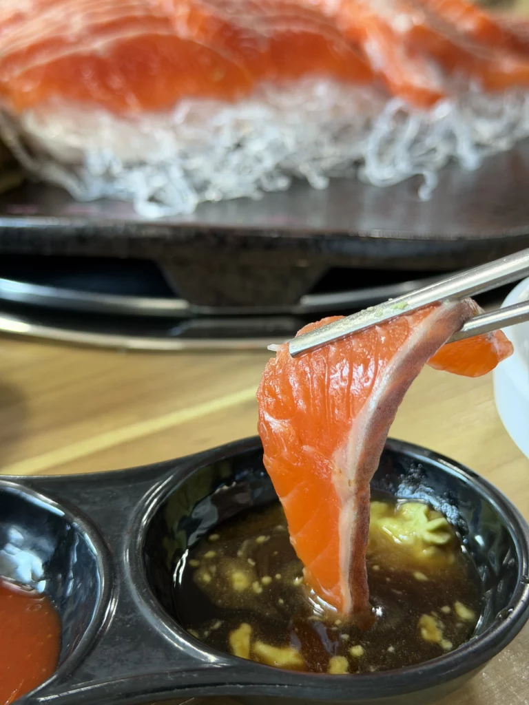 Chopsticks dipping a fresh slice of trout sashimi (송어회) into wasabi soy sauce (와사비 간장) — the Japanese-influenced dipping option served alongside choganjang at Vivaldi Songeogol in Gangwon Province.