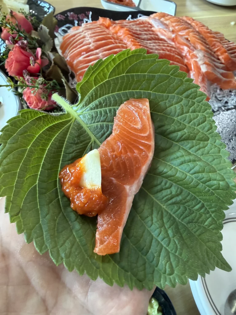 A slice of fresh trout sashimi (송어회) placed on a Korean perilla leaf (깻잎) with choganjang and garlic, ready to be wrapped and eaten ssam-style at Vivaldi Songeogol.