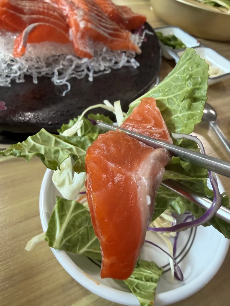Chopsticks placing a fresh slice of trout sashimi (송어회) over a bowl of shredded vegetables — the Korean technique of eating freshwater fish with a crunchy vegetable mix and choganjang at Vivaldi Songeogol.