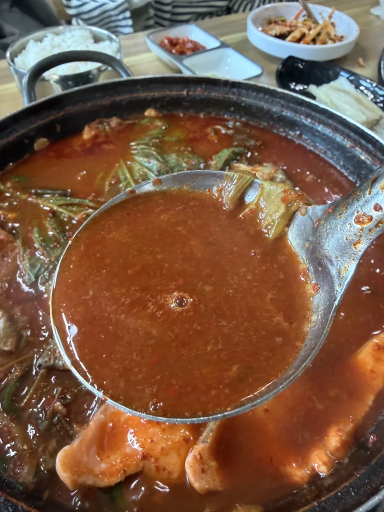 A ladle lifting the rich, deep red gochugaru broth of trout maeuntang (매운탕) — the intensely flavored spicy fish stew made from fresh trout bones and head that concludes every Korean trout sashimi (송어회) meal at Vivaldi Songeogol.