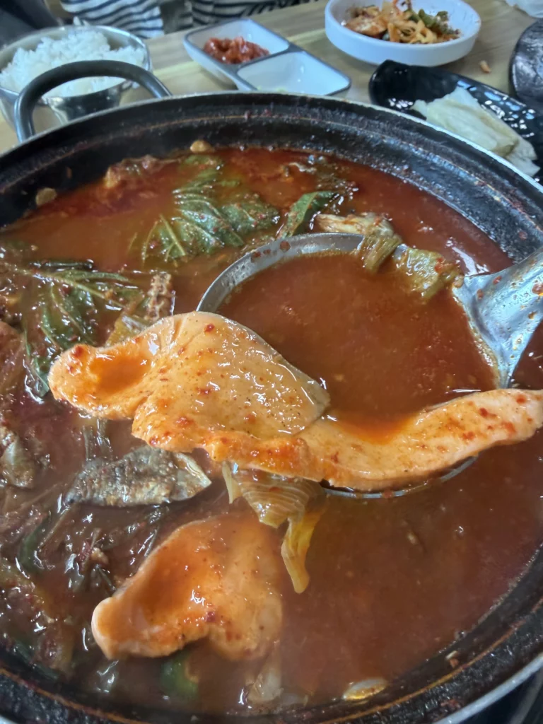 A ladle lifting a piece of trout head from bubbling maeuntang (매운탕) at Vivaldi Songeogol — the spicy fish stew made from leftover trout bones and head that traditionally closes a Korean trout sashimi (송어회) meal in Gangwon Province.