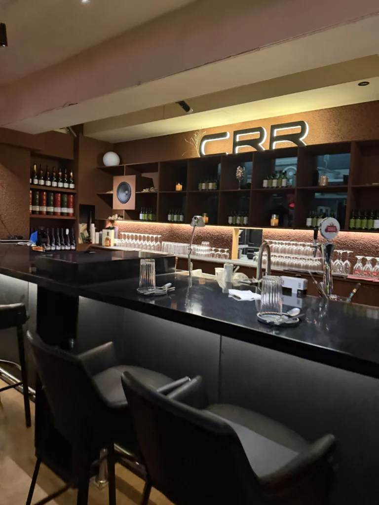 Bar counter area at Charari in Sinsa-dong Seoul, featuring a black countertop, backlit shelves stocked with Korean traditional spirits and wine, hanging glassware, and a illuminated 