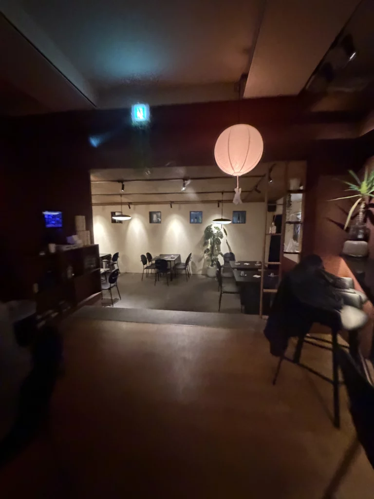 View from the main dining room into the secondary dining area at Charari in Sinsa-dong Seoul, with a round paper lantern in the foreground, warm spotlighting, framed wall art, and a large indoor plant — highlighting the restaurant's intimate two-room layout.