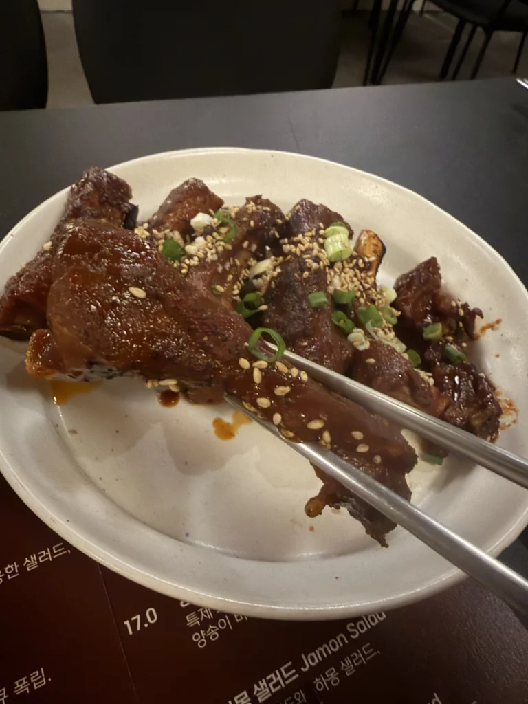 Tongs lifting a glazed piece of 불등갈비 (fire galbi) from a white plate at Charari in Sinsa-dong, showcasing the caramelized marinade, sesame seeds, and tender back rib texture up close.