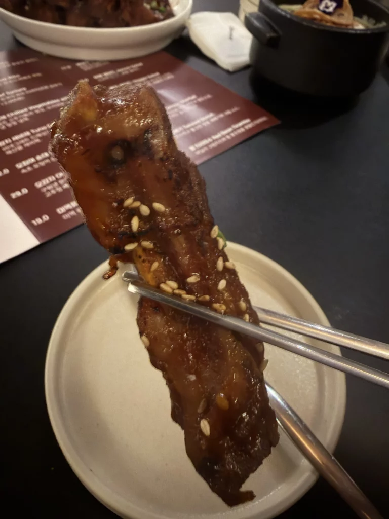 Close-up of a single fire-marinated galbi rib held by tongs at Charari in Sinsa-dong, revealing the deeply lacquered, caramelized exterior and sesame seed topping of the signature 불등갈비.