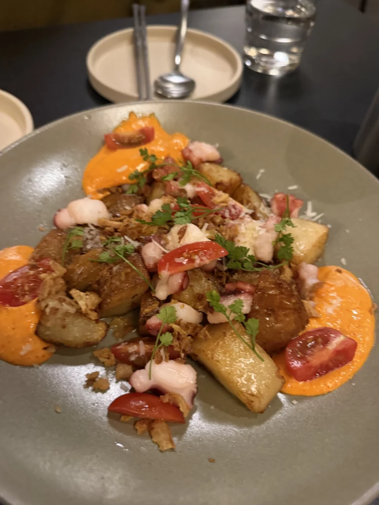 Charari's 오감자 (Ogamja) side dish plated on a grey bowl, showing roasted potato pieces topped with cherry tomatoes, octopus, crispy garnish, fresh herbs, and orange sauce dollops — a contemporary Korean accompaniment to the fire galbi menu.