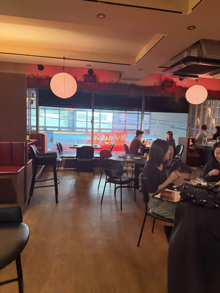 Daytime interior of Charari restaurant in Sinsa-dong Seoul, featuring white paper lanterns, dark leather seating, and large windows overlooking the street, with diners seated at tables in a modern Korean galbi dining setting.
