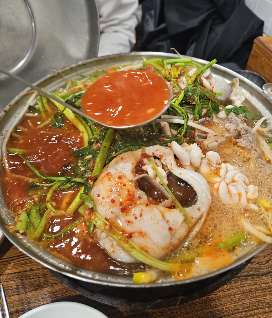 Spicy Won Daegu-tang Cod-fish Hot Pot with cod fish slices, vegetables, and red chili broth being poured into the bubbling Korean seafood hot pot.
