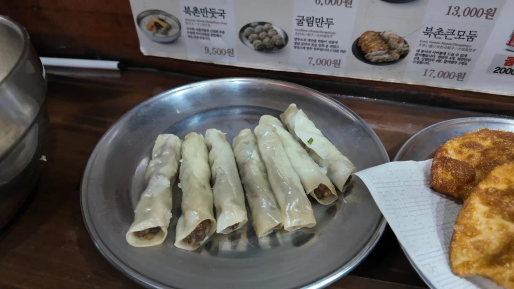 Bukchon Son Mandu thin-skin mandu rolls displayed on a plate with menu board showing prices in the background.