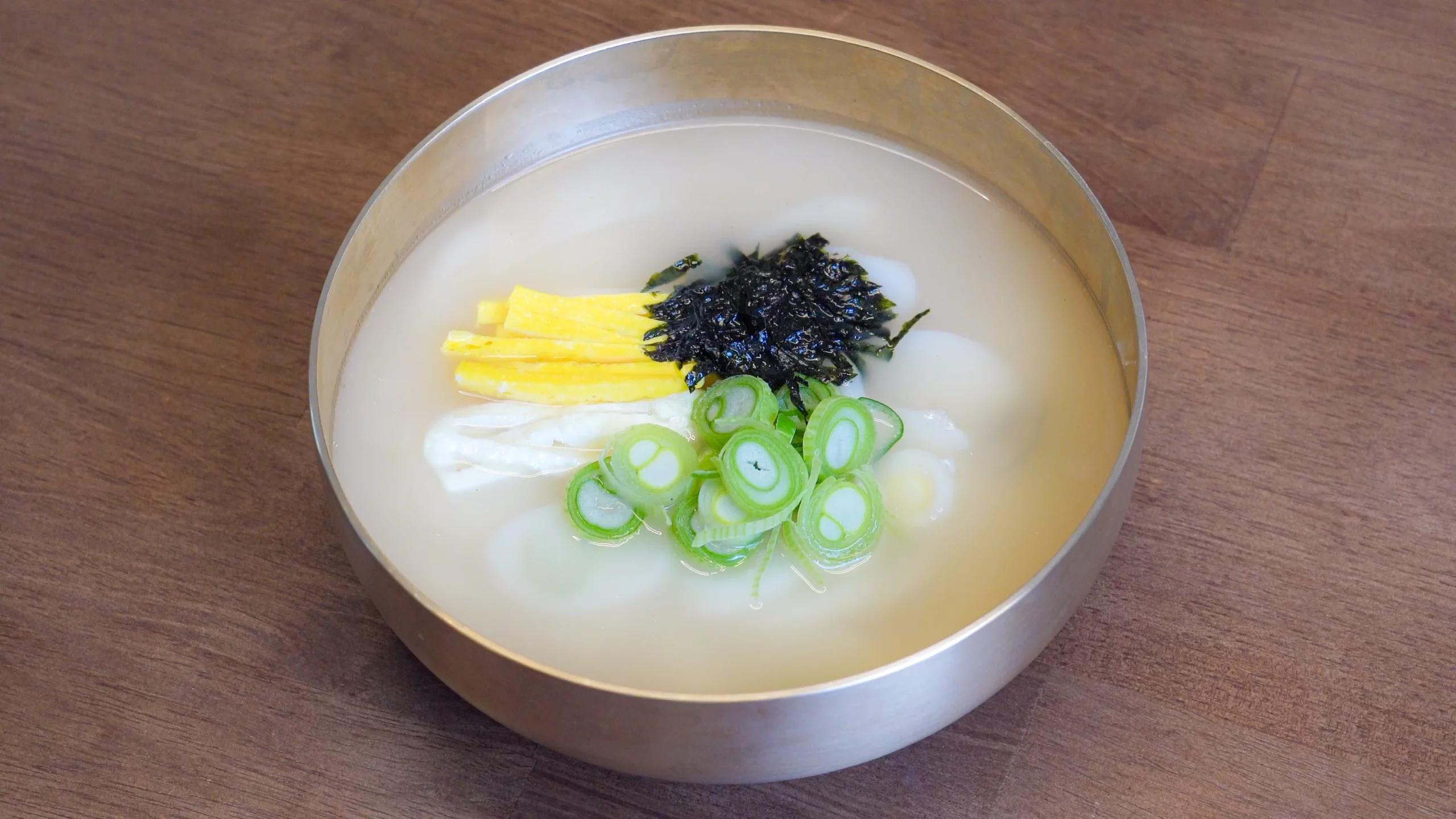 Top-down view of homemade tteokguk (Korean rice cake soup) in a brass bowl with chewy oval rice cakes in clear broth, garnished with egg yolk and white jidan, crushed roasted seaweed, and green onion rings