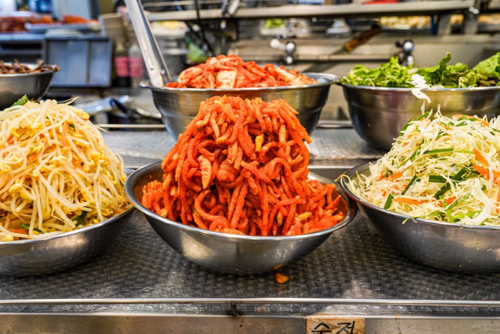 Multiple stainless steel bowls of prepared bibimbap toppings at Gabose Bibimbap including spicy radish kimchi, seasoned bean sprouts, and shredded cabbage salad